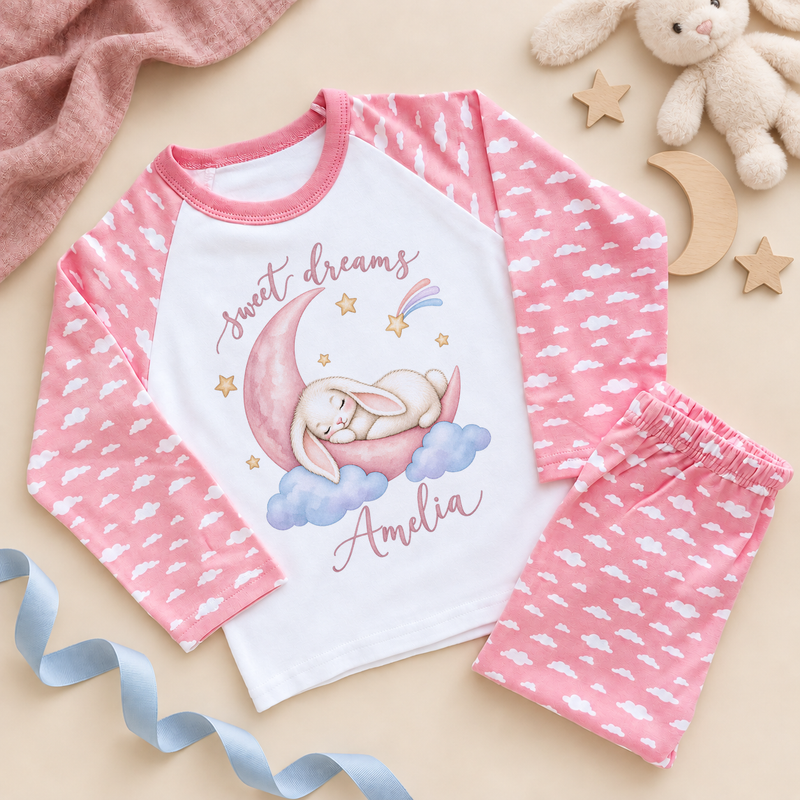 Personalised pink cloud pyjamas: a cream sleeping bunny on a pink crescent moon with sweet dreams script, gold stars and an example name in pink below the rabbit, on a white raglan top with matching pink cloud print bottoms